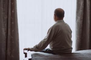 picture of an elderly man sitting on his bed in shadow and looking out of his window